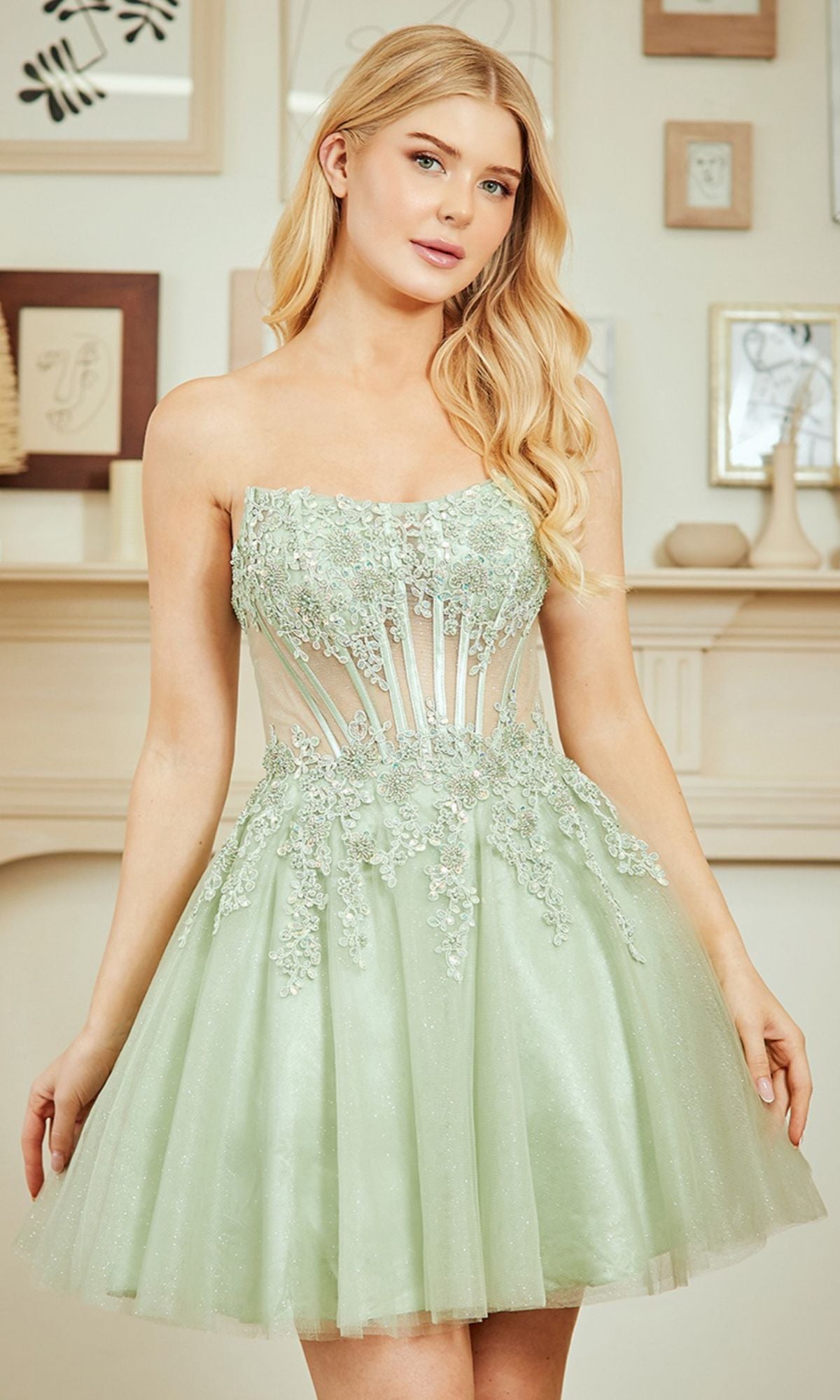 A woman wears the Amelia Couture 9083S Strapless Short Party Dress, a light green corset-style fit with floral embroidery, standing indoors by a decorated wall—ideal for a stylish homecoming look.